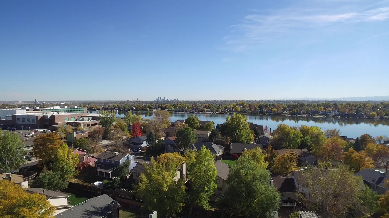 Opening aerial frame over Hidden Lake, Colorado, with residential roofs and autumn trees in the foreground, a calm lake behind them, a brick building at left, distant skyline on the horizon, and no visible people.