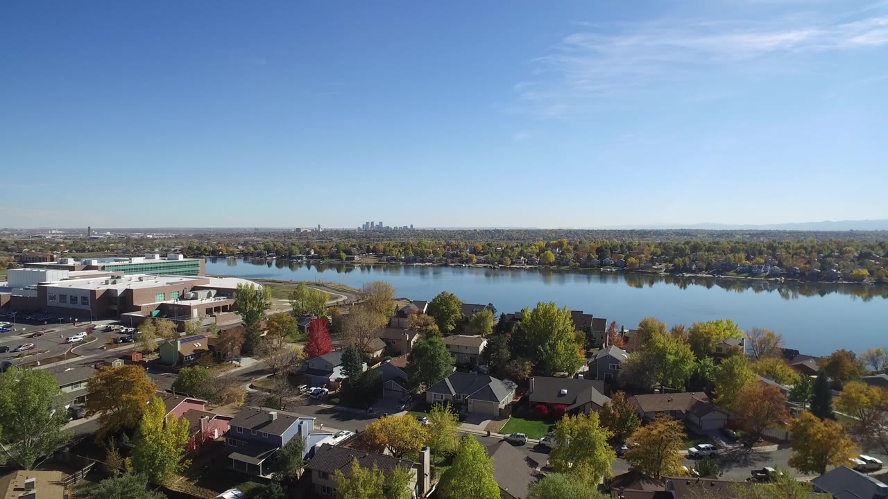 Opening aerial frame of Hidden Lake, Colorado with foreground homes, autumn trees, a large building at left, the lake across the middle, distant skyline, and no visible people.
