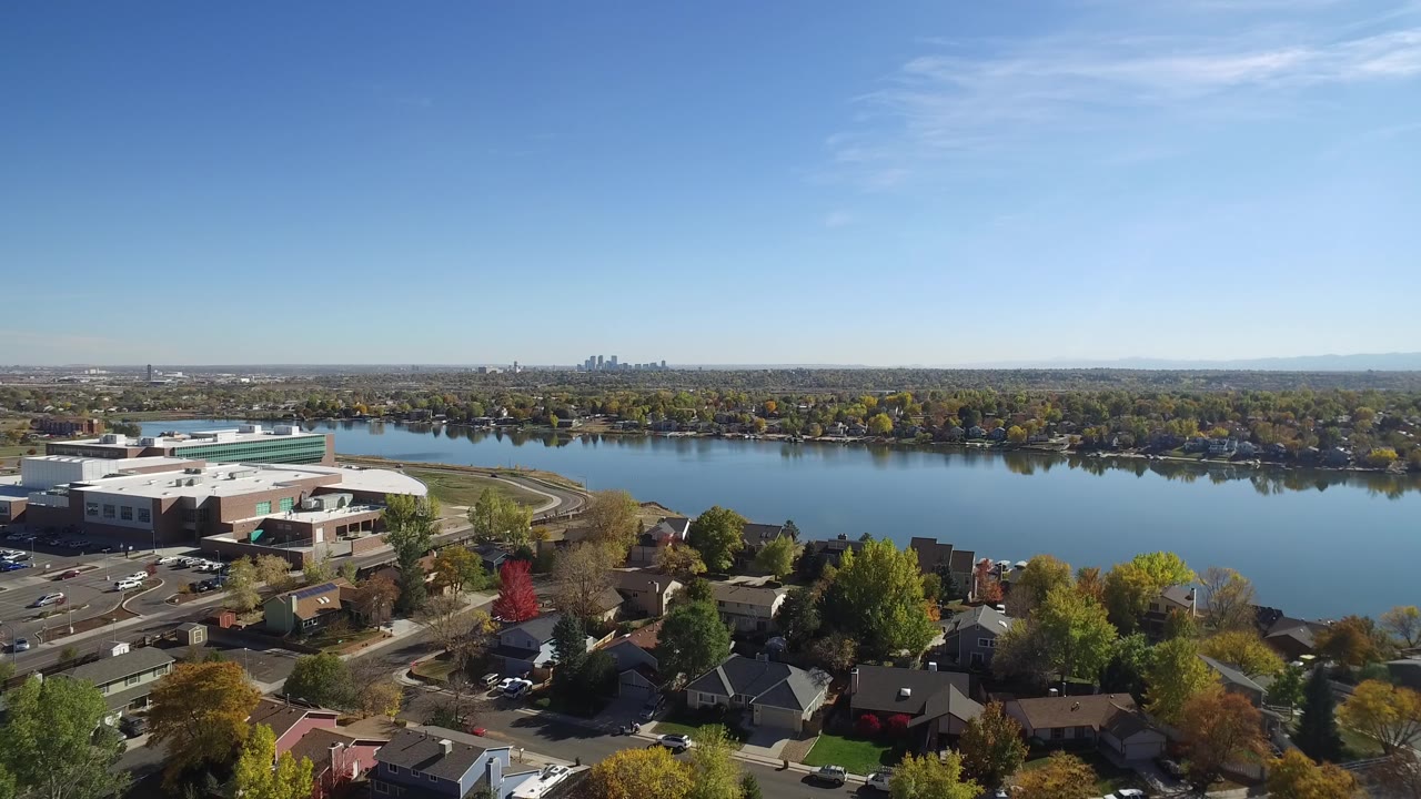 Aerial view of a lake, homes, streets, and a distant skyline under blue sky.