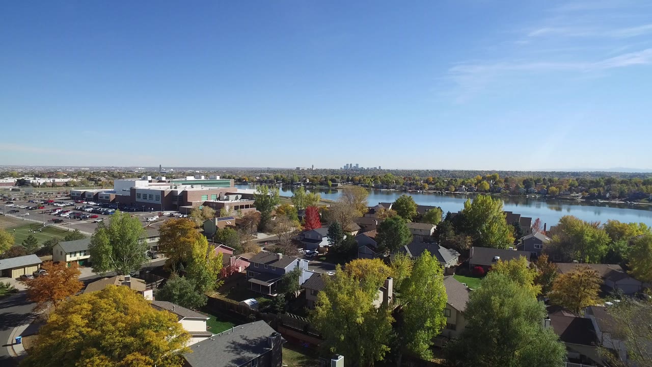 Aerial view of homes, trees, a lake, and distant buildings under a clear sky.