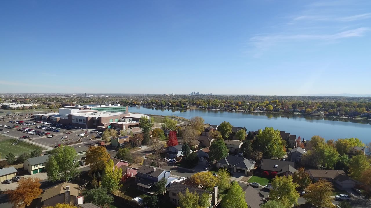 Aerial view of a school, parking lots, homes, trees, and a lake under blue sky.