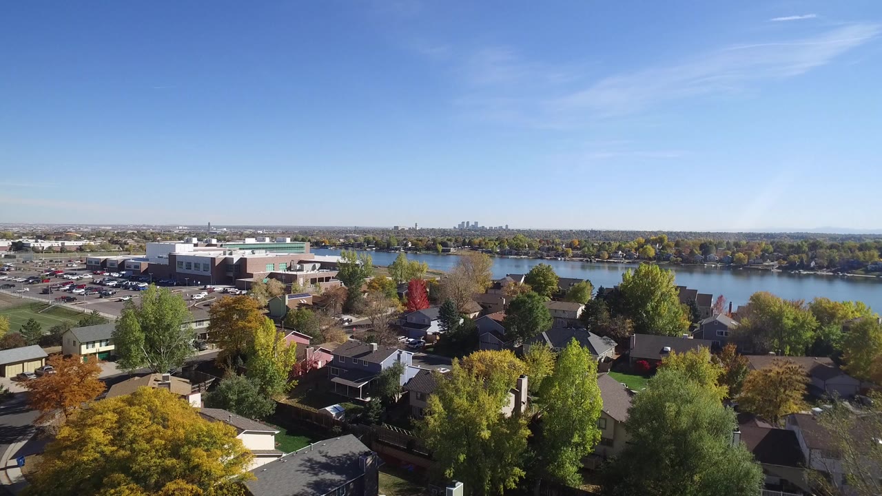 Aerial view over homes, autumn trees, a lake, parking area, and distant skyline.