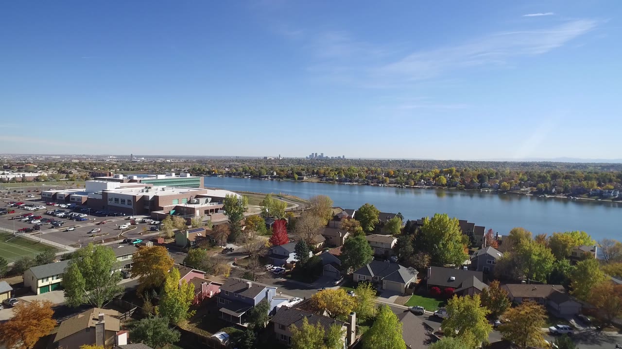 Opening aerial frame near Hidden Lake, Colorado, showing residential rooftops and trees in the foreground, a large pale building and parking lot to the left, blue water across the middle, and no visible people.