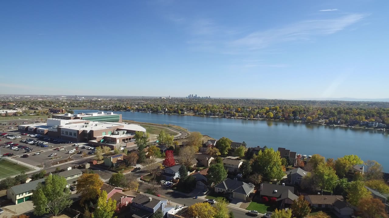 Opening aerial frame over Hidden Lake, Colorado, with foreground homes and trees, a large white building and parking lot at left, blue lake water in the midground, distant skyline, and no visible people.