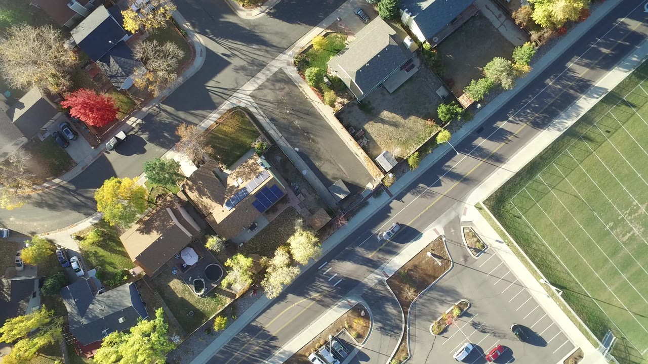 Overhead view of suburban houses, streets, parking spaces, trees, and a green athletic field.