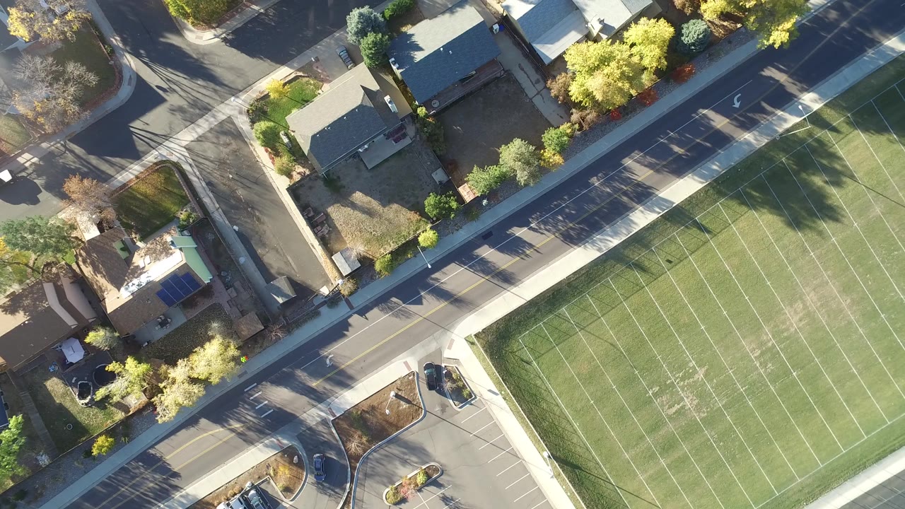 Overhead frame in Hidden Lake, Colorado with the football field on the right, a parking lot at lower left, houses along the upper edge, and tiny distant figures or dark marks on the turf.