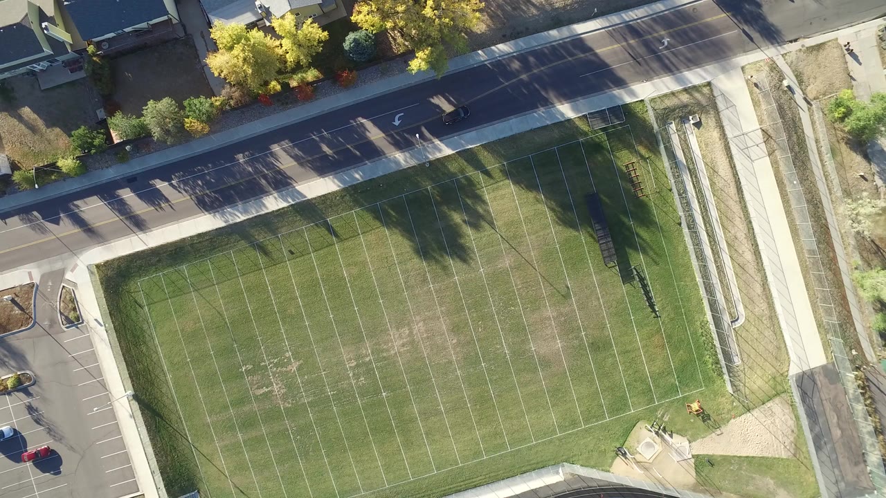 Overhead aerial frame of an empty grassy sports field in Hidden Lake, Colorado, with roadways, trees, sidewalks, fencing, parked cars, and no visible people.