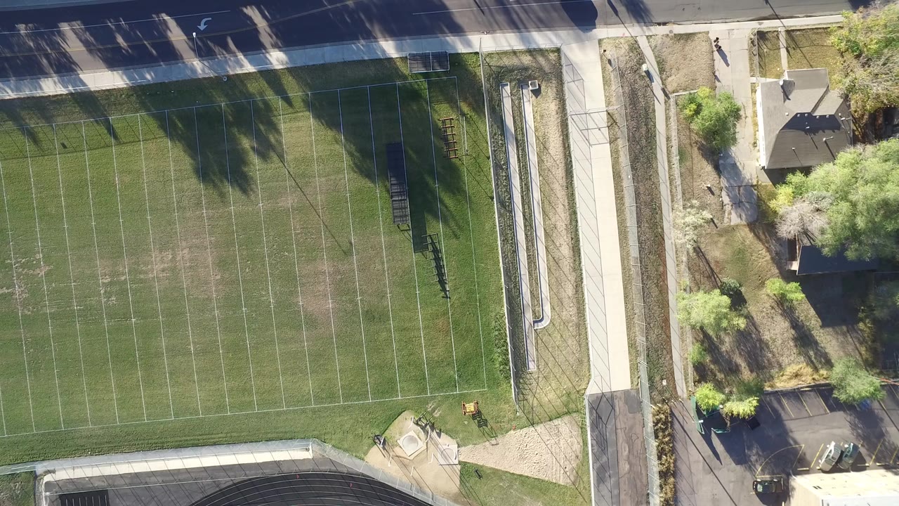 Top-down view of a marked green football field beside paths, road, trees, and buildings.