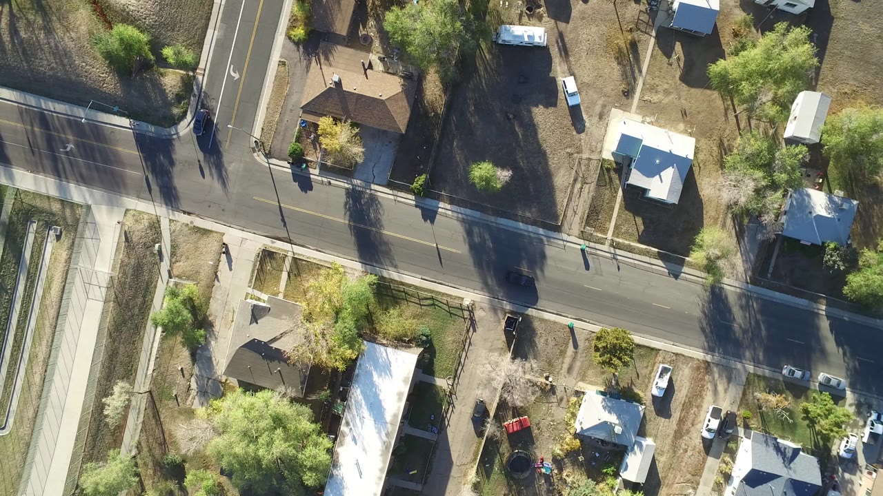 Overhead drone frame in Hidden Lake, Colorado showing a main road crossing below smaller lots, light-roofed homes, parked vehicles, trees, and no visible people.
