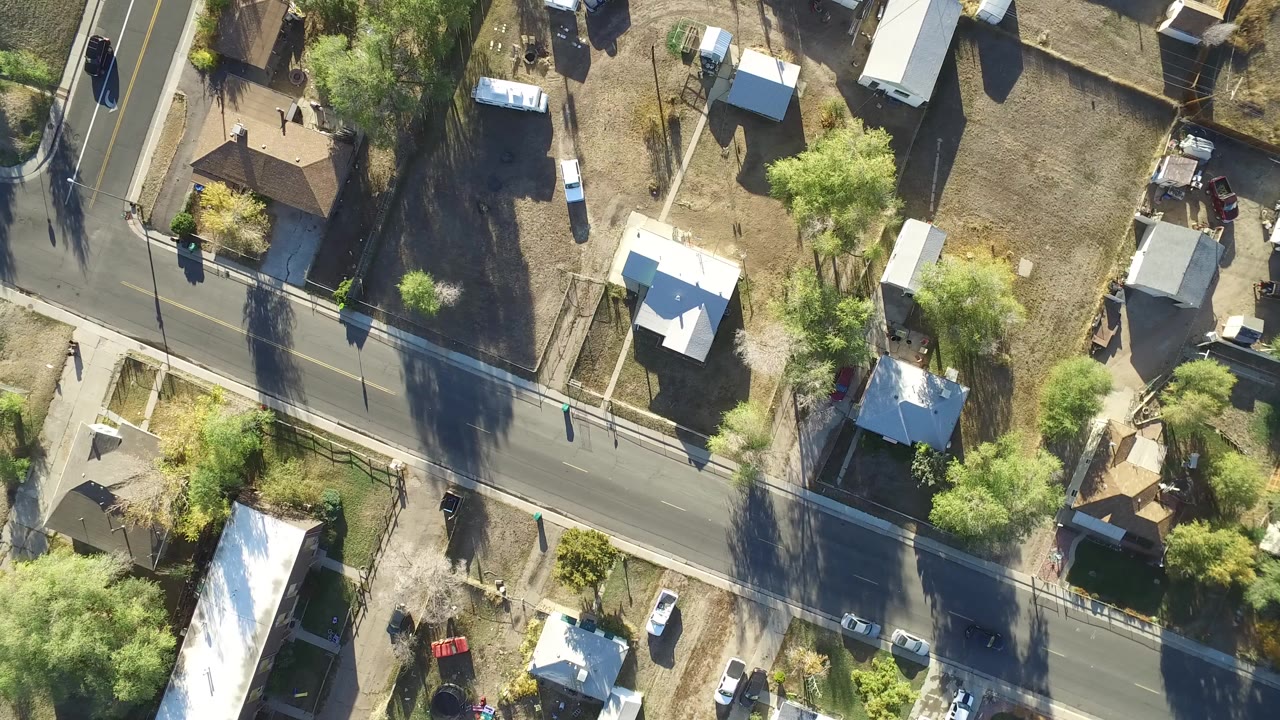 Overhead drone frame of Hidden Lake, Colorado showing a diagonal residential road, small homes, dry yards, parked cars, scattered trees, and tiny people near the upper-left property.