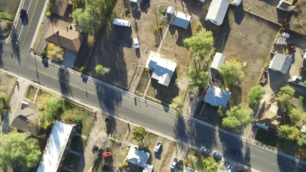 Aerial frame over Hidden Lake, Colorado with a paved road running across the lower half, white-roofed houses, open brown yards, trees casting long shadows, and parked vehicles.