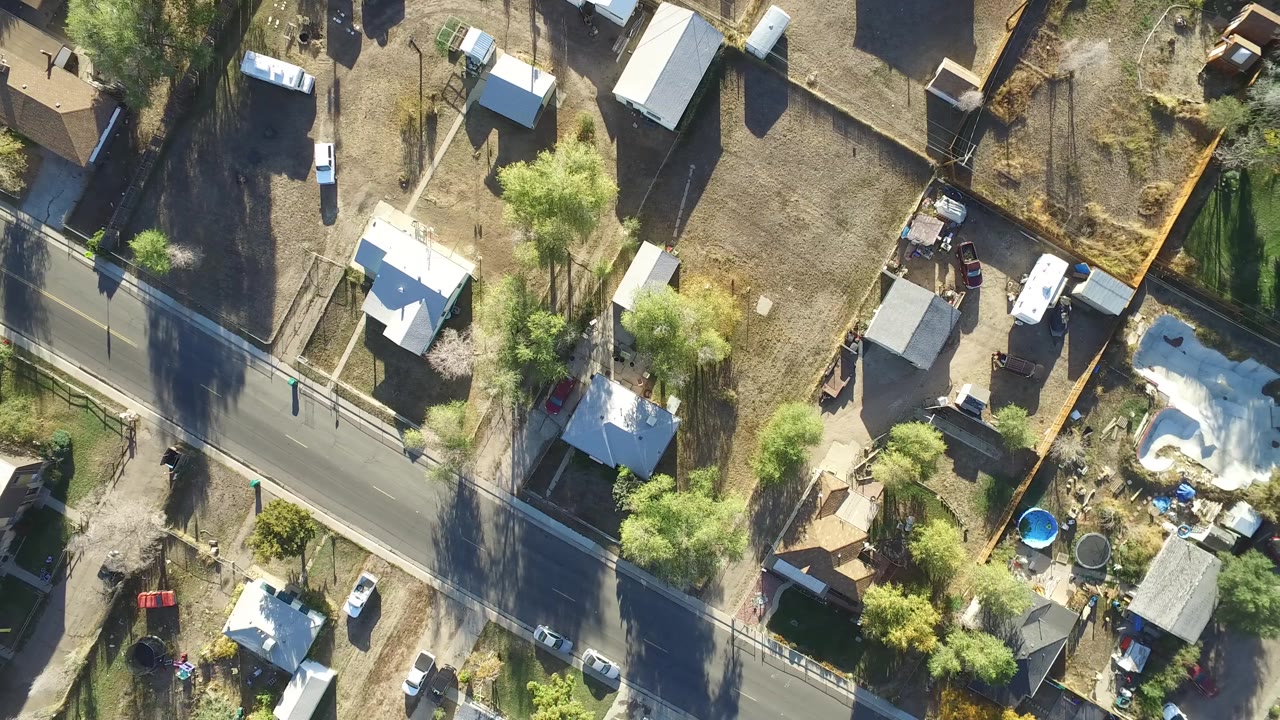 Aerial overhead frame in Hidden Lake, Colorado with the road running diagonally past fenced lots, light-roofed houses, dry ground, trees, parked vehicles, and backyard pools, with no visible people.