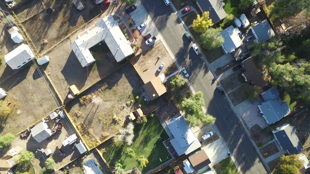 Overhead drone frame in Hidden Lake, Colorado showing light-roofed homes, fenced yards, a residential street, parked cars, dry lots, green trees, and no visible people.