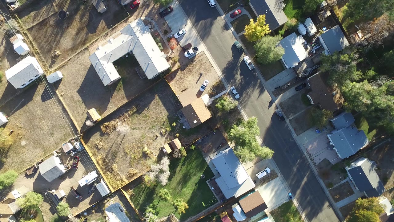 Aerial frame over Hidden Lake, Colorado with rooftops, fenced backyards, a paved road, parked vehicles, green lawns, dry ground, tree shadows, and no visible people.