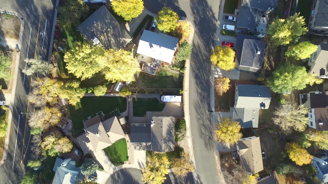 Top-down aerial frame over Hidden Lake, Colorado showing detached houses, green lawns, yellow-green trees, residential roads, and parked cars, with no clearly identifiable faces visible.