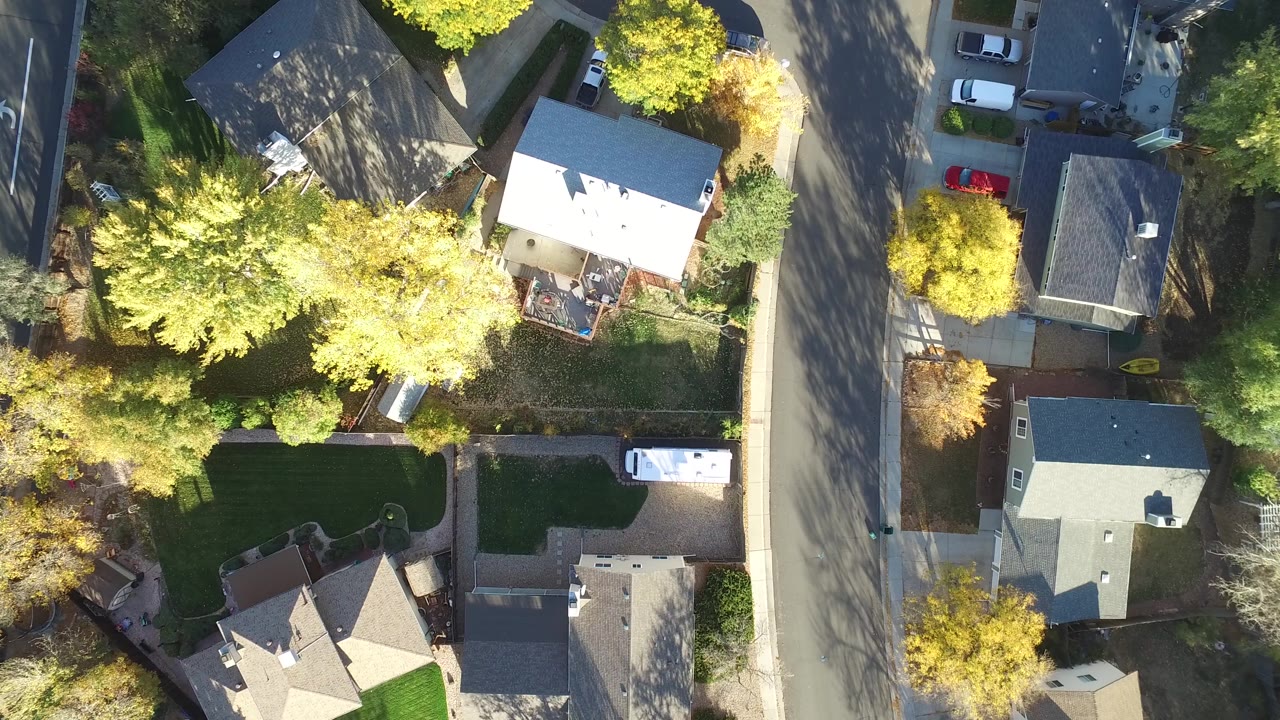 Aerial view in Hidden Lake, Colorado with a bright white-roofed house near the center, a curved street, fenced yards, parked vehicles, trees, and no clearly visible people.