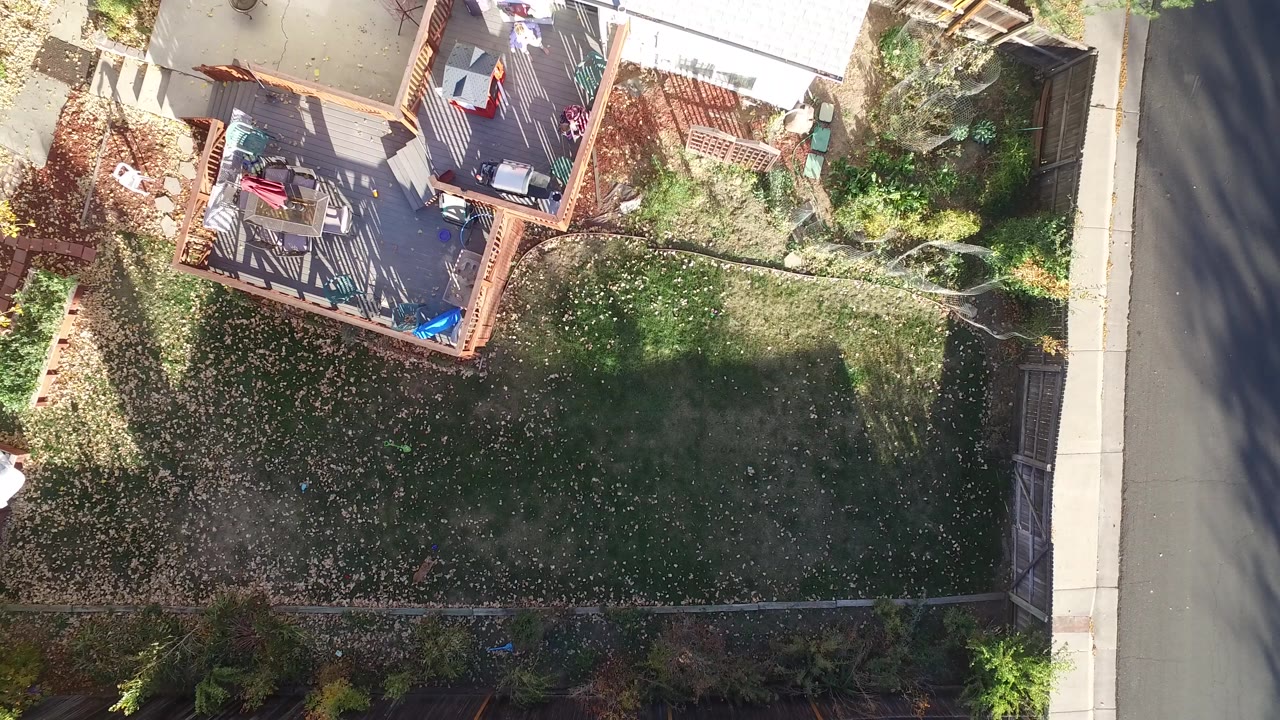 Overhead drone frame from Hidden Lake, Colorado showing a fenced backyard with a wooden deck at upper left, a leaf-strewn lawn below, and a road edge on the right.