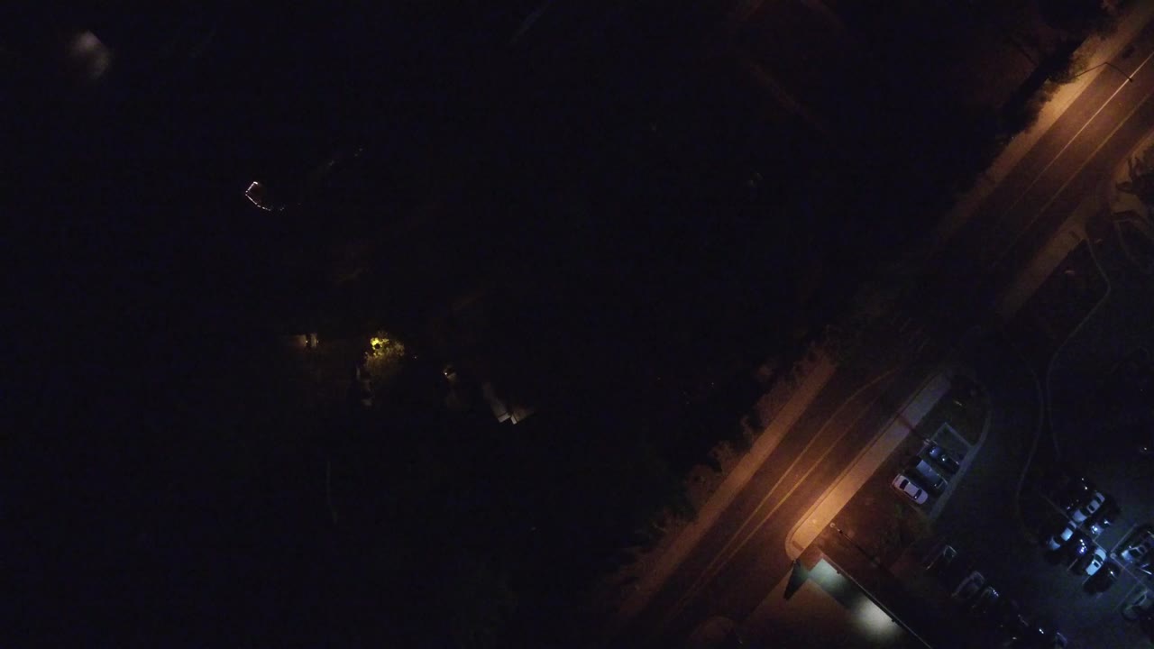 Overhead night frame from Hidden Lake, Colorado showing a diagonal orange-lit road, a small cool-blue parking lot, dark surrounding ground, and no visible people.