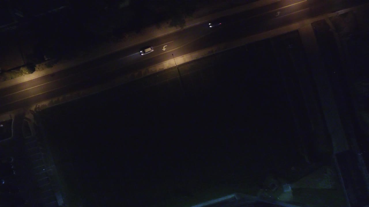 Overhead night view of a lit road with a few cars and dark buildings below.