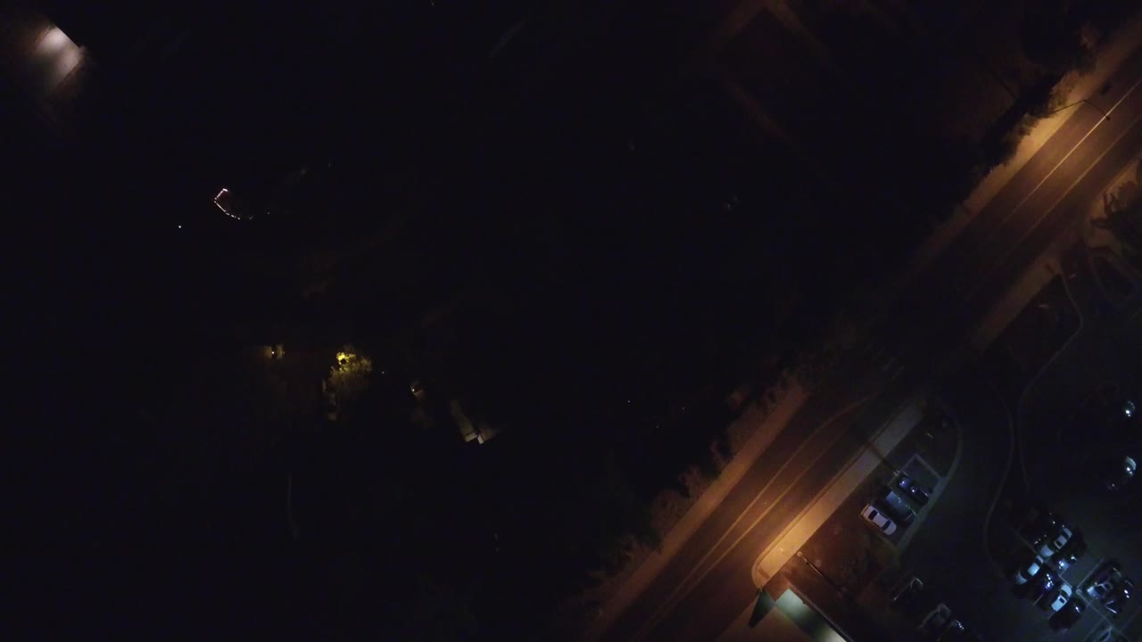 Overhead night frame in Hidden Lake, Colorado showing a diagonal lit street, a small parking area with cars, surrounding darkness, and no visible people.