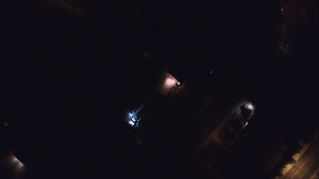 Very dark aerial view with a lit road edge and scattered building lights.