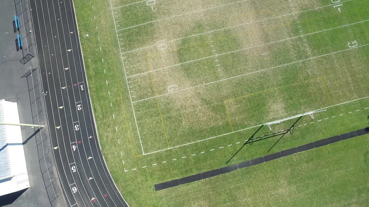 Overhead view in Hidden Lake, Colorado, showing a green football field with white yard lines, a black running track on the left, nearby roof edges, and no visible people.
