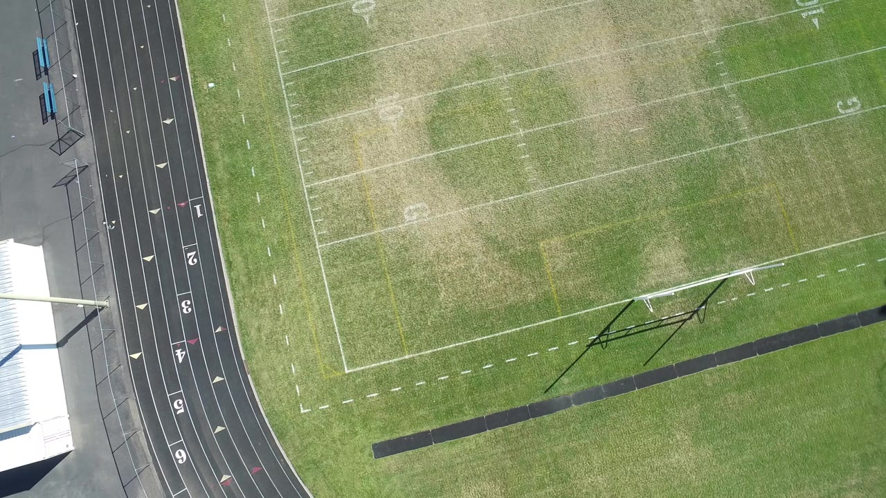 High overhead frame in Hidden Lake, Colorado, with the football field angled across the view, the black track at left, a small goal or practice frame shadow, and no visible people.