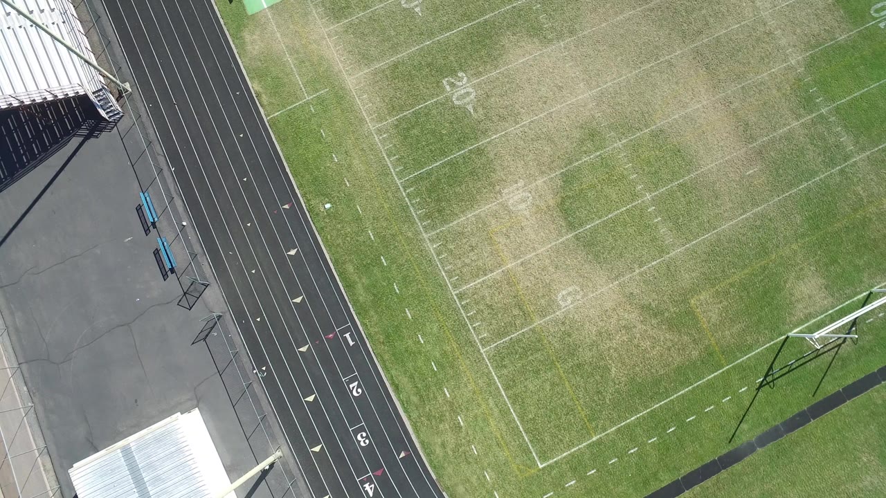 Overhead frame in Hidden Lake, Colorado showing a football field beside a black running track, metal bleachers, pavement, and no visible people.
