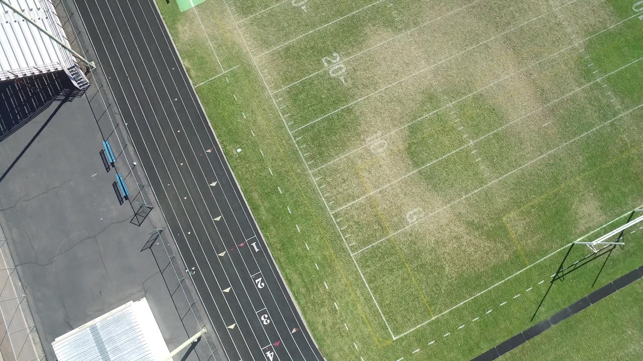 Aerial frame over the Hidden Lake, Colorado stadium with track lanes along the left side, faint field numbers on patchy turf, and no visible people.