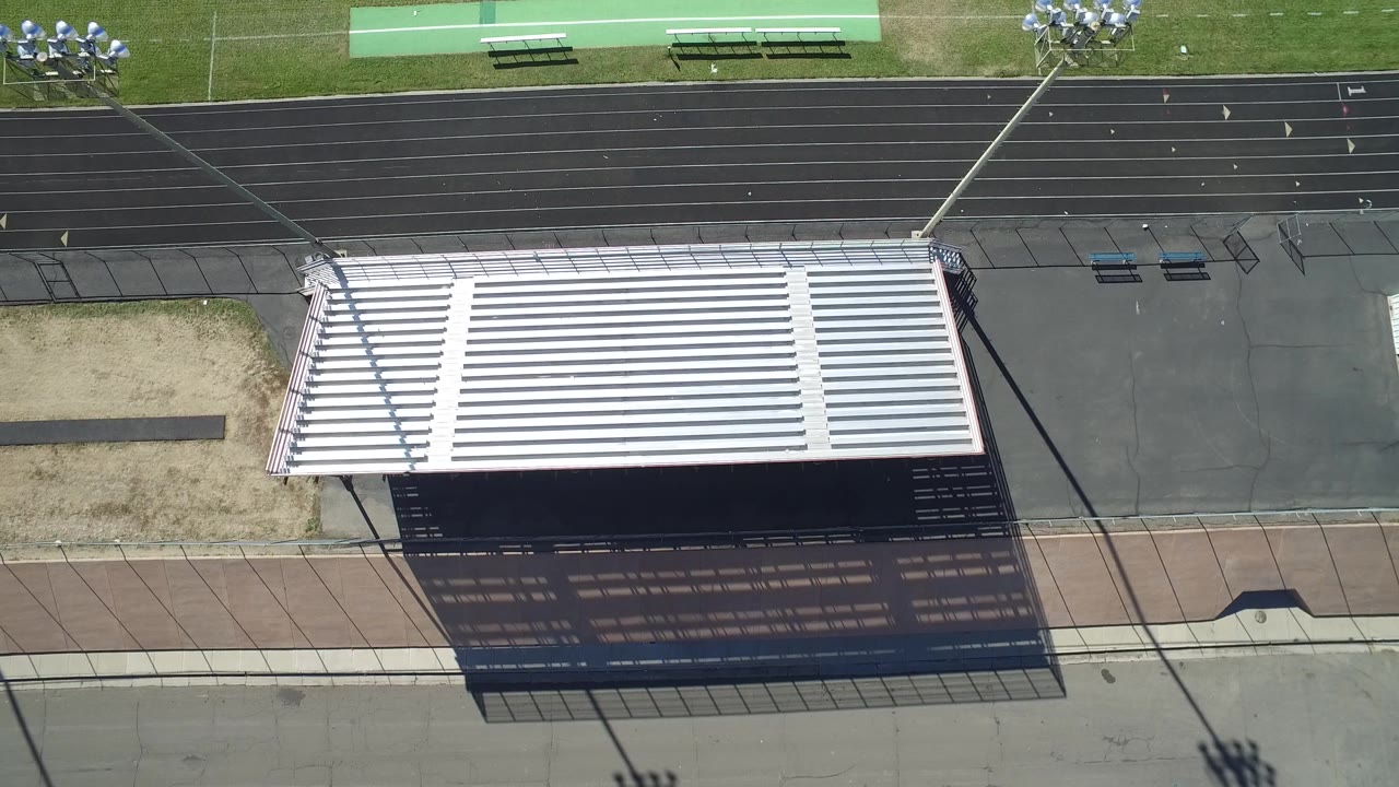 High aerial frame in Hidden Lake, Colorado with the white bleachers centered, their shadow falling across pavement, the black track above, and a small group of visible people at the top right.