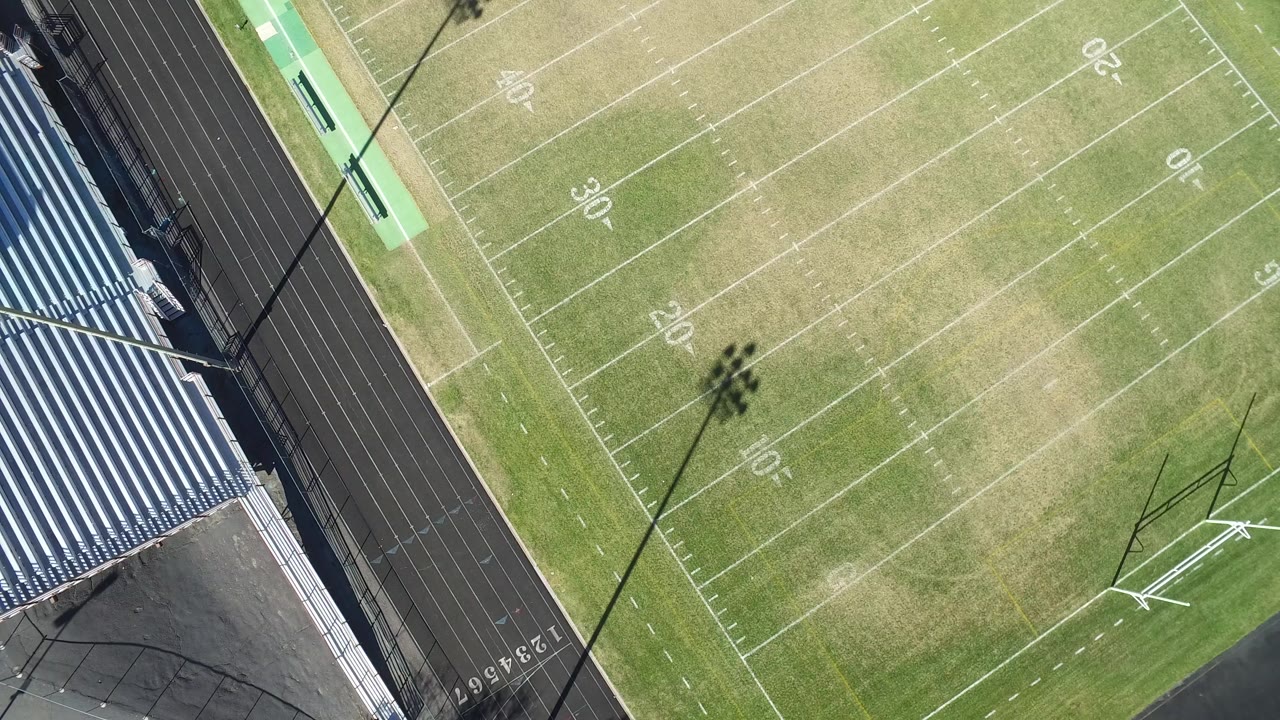 Overhead drone frame in Hidden Lake, Colorado showing football yard lines, part of a black running track, blue bleachers, and long light pole shadows with no visible people.