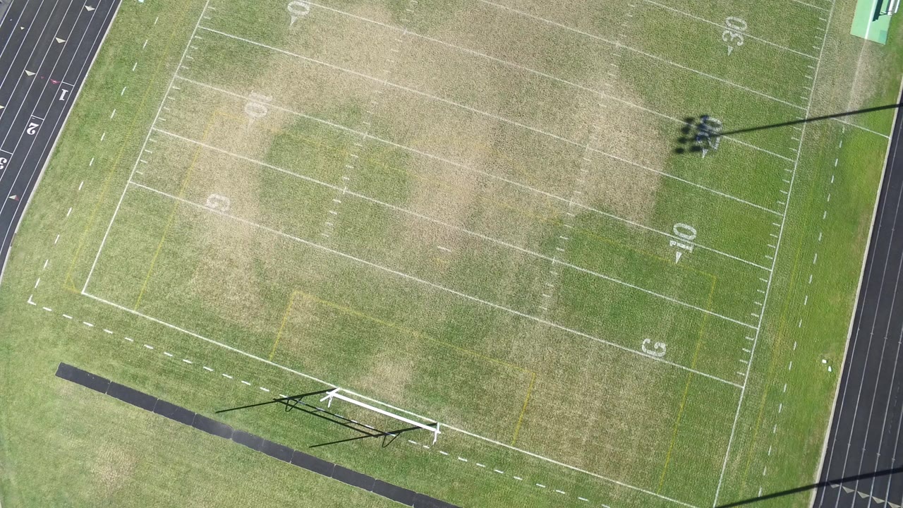 High aerial view of a football field in Hidden Lake, Colorado, with white yard lines, worn grass patches, a goalpost shadow, and no visible people.