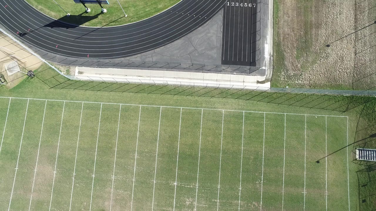 Overhead aerial frame in Hidden Lake, Colorado showing the upper section of an empty football field, the curve of a dark running track, bright daylight, and no visible people.