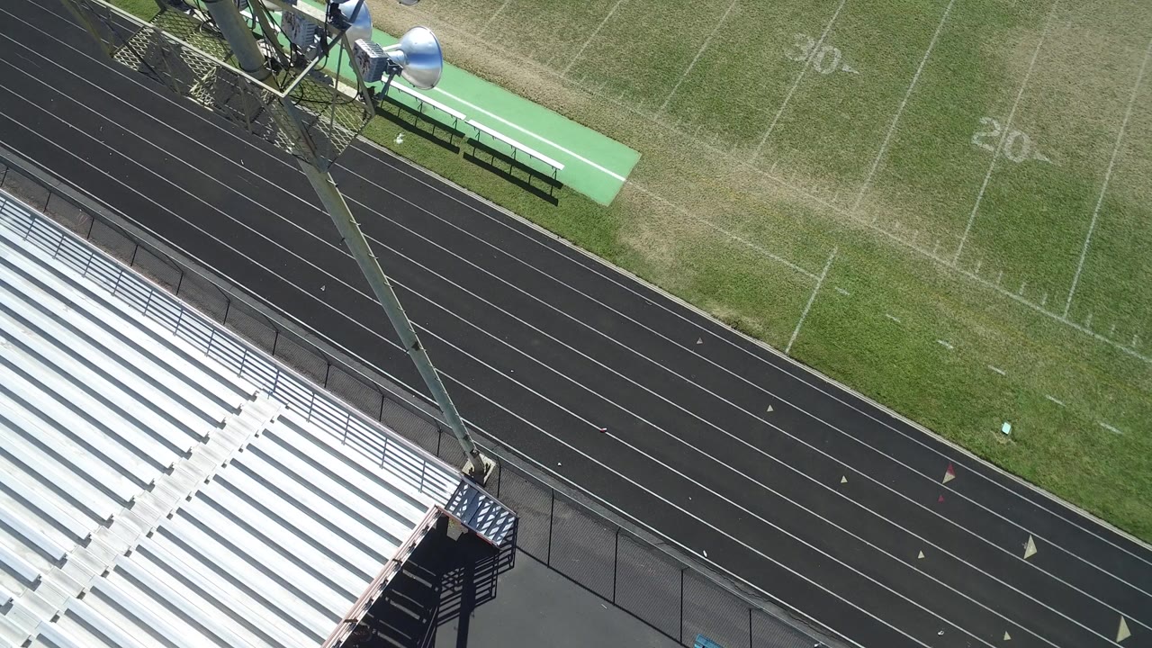 Aerial frame over a Hidden Lake, Colorado stadium showing white bleachers at lower left, a black running track, green field markings, benches, light poles, and no visible people.
