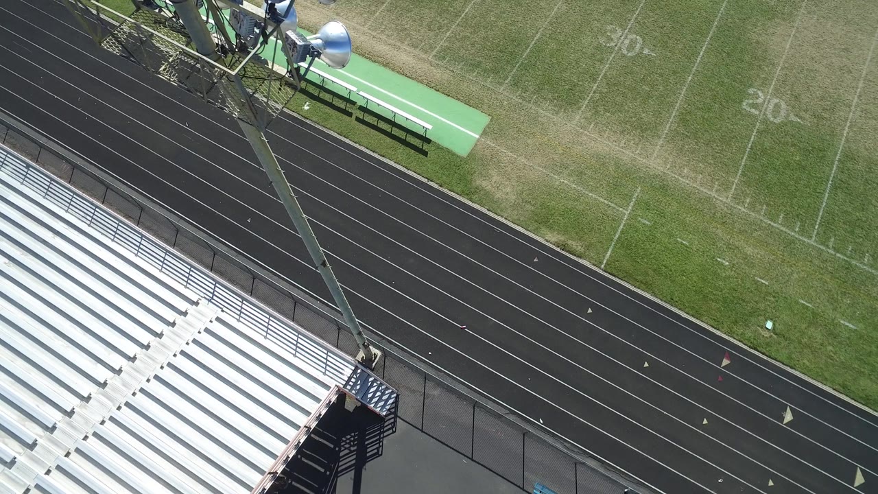 A slightly shifted aerial frame in Hidden Lake, Colorado shows empty white bleachers, multiple black track lanes, green turf with faint yard numbers, benches, and no visible people.