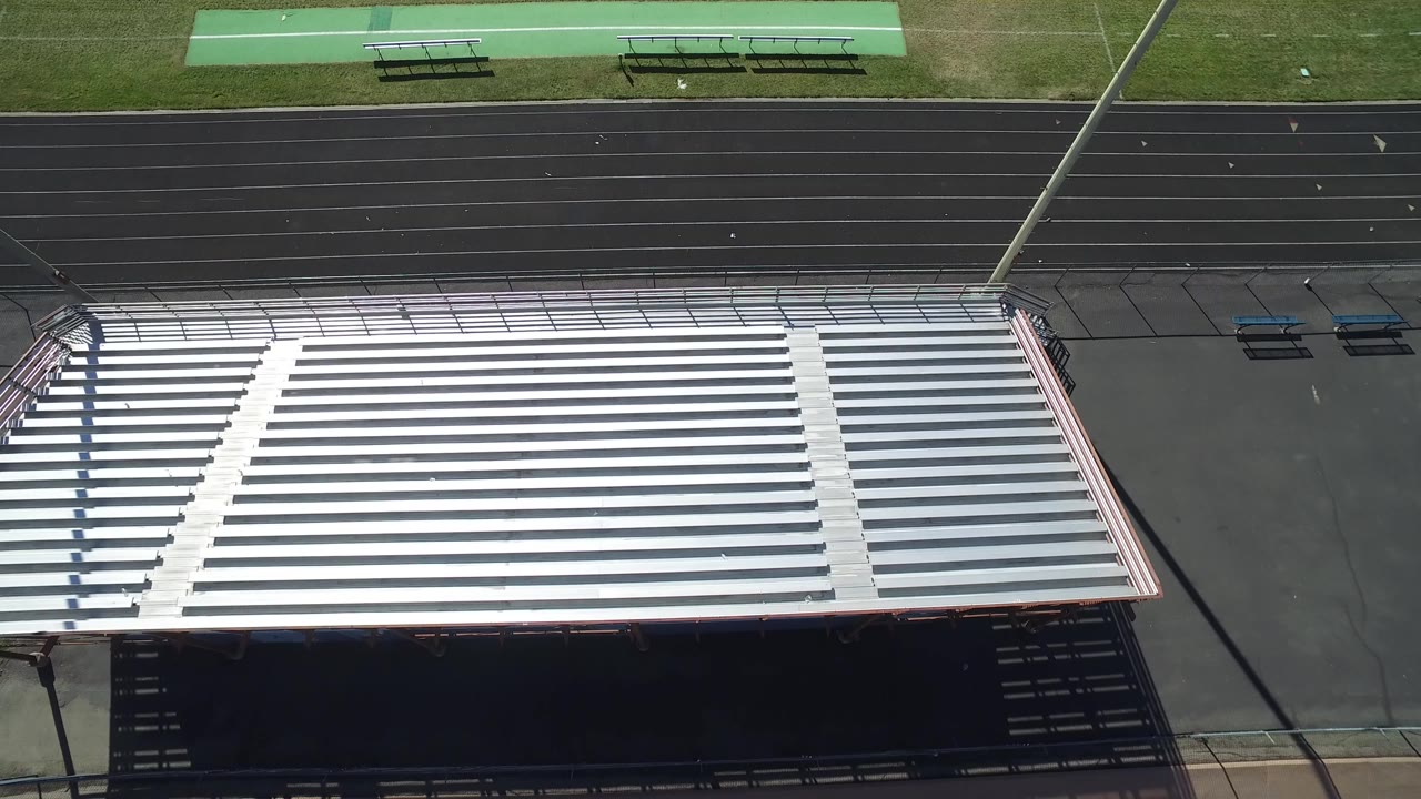 High aerial frame over silver bleachers, a black running track, and green field edge in Hidden Lake, Colorado, with no visible people.