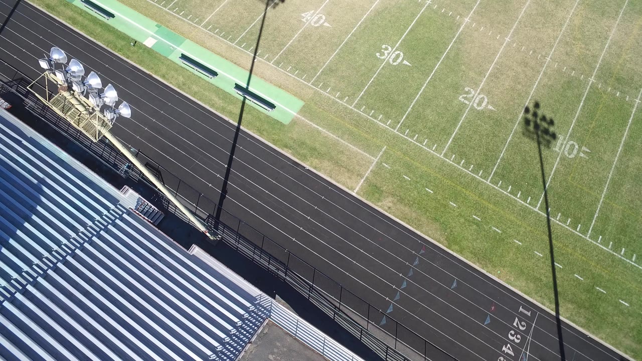 Overhead frame from Hidden Lake, Colorado showing striped bleachers beside a black running track and a green football field with yard numbers, with no visible people.