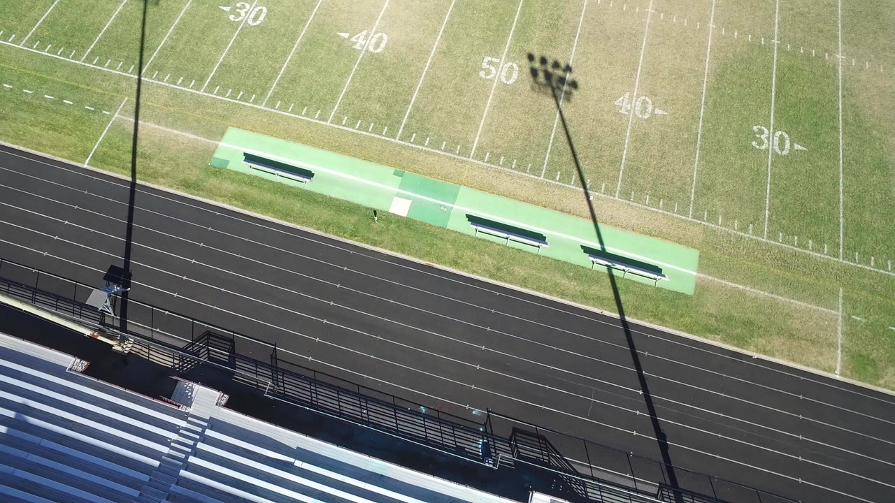 Overhead frame in Hidden Lake, Colorado showing an empty football field above a black running track, with bleachers at the bottom edge, sideline benches, a stadium light pole shadow, and no visible people.