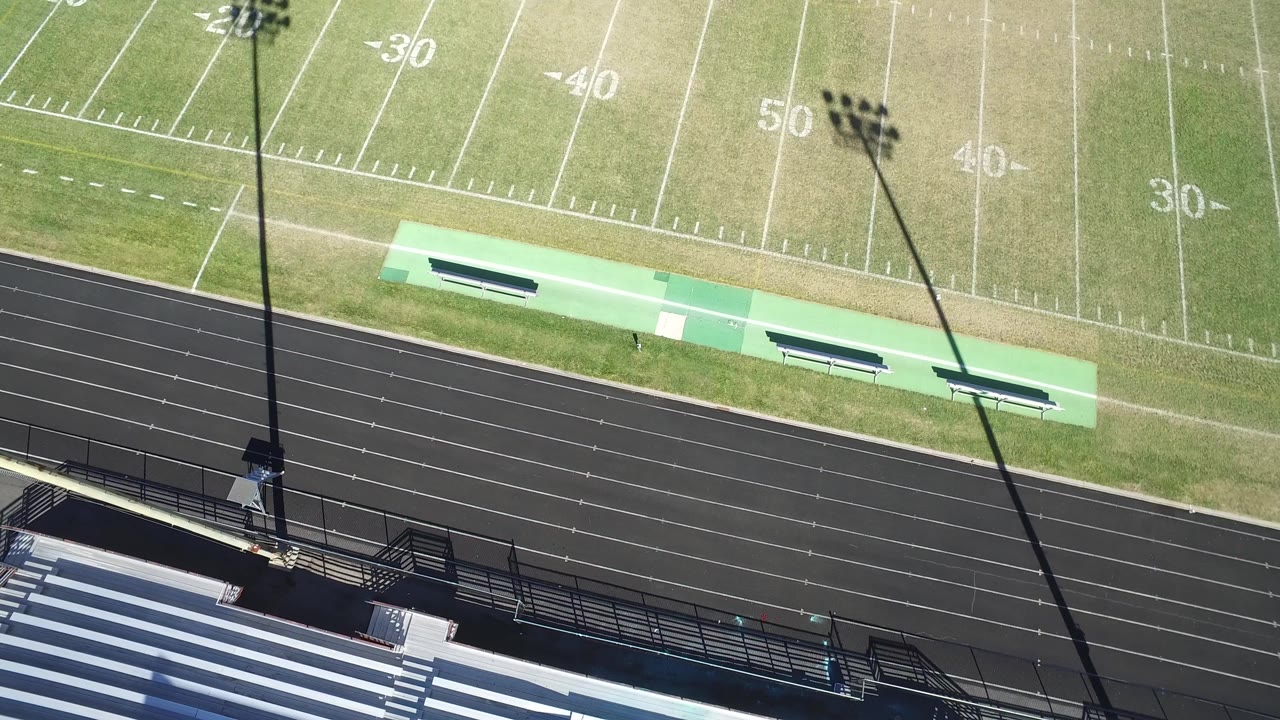 Drone-style overhead of football yard lines, track, benches, and bleachers.