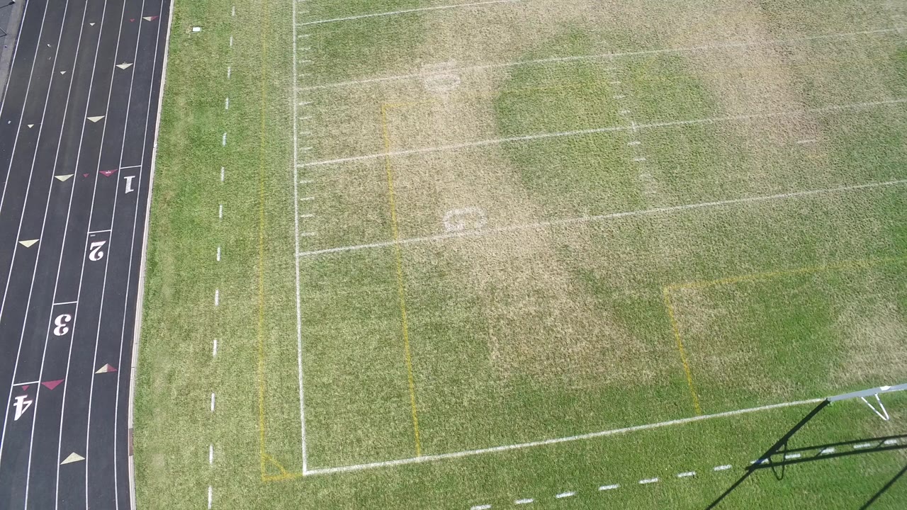 Overhead view in Hidden Lake, Colorado, showing a green football field with worn grass patches beside a black running track, with no visible people.