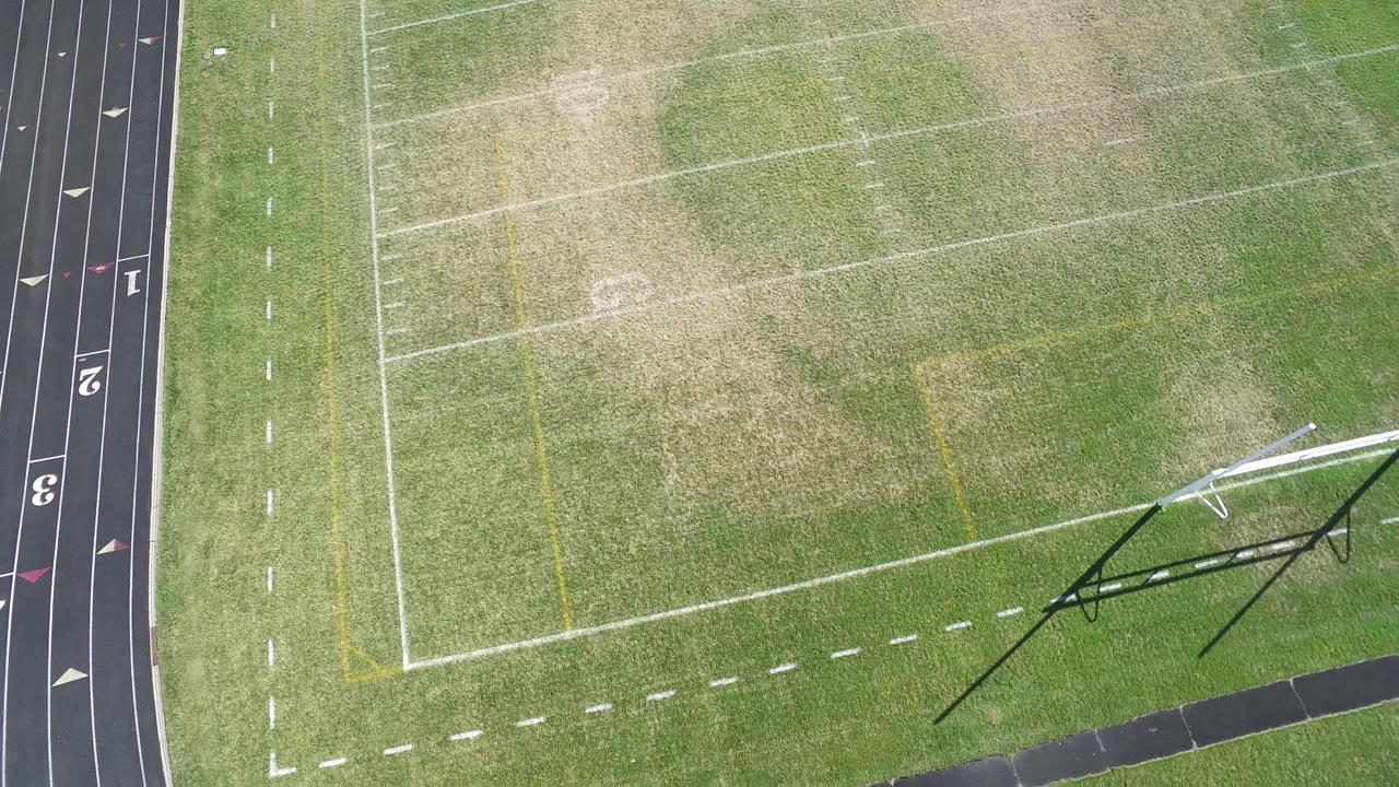 Aerial frame from Hidden Lake, Colorado showing worn grass, white football markings, yellow field lines, black track lanes, and the goalpost area with no visible people.
