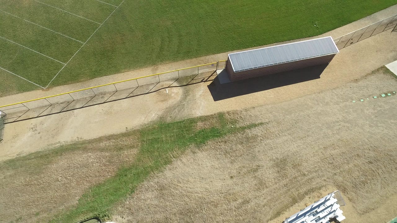 Aerial frame from Hidden Lake, Colorado showing a football field edge, diagonal fence, tan dirt area, green turf, a small metal-roofed building, and no visible people.
