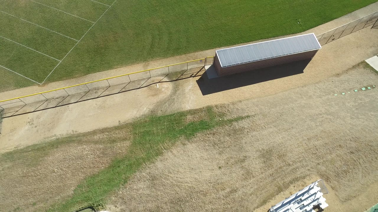 Drone-like overhead view of football field grass, fence shadows, dirt, and a small utility building.