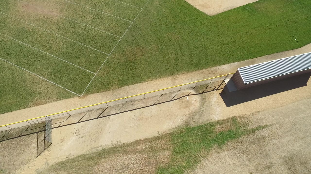Aerial view of a fenced grass field beside a gravel path and small roofed structure.