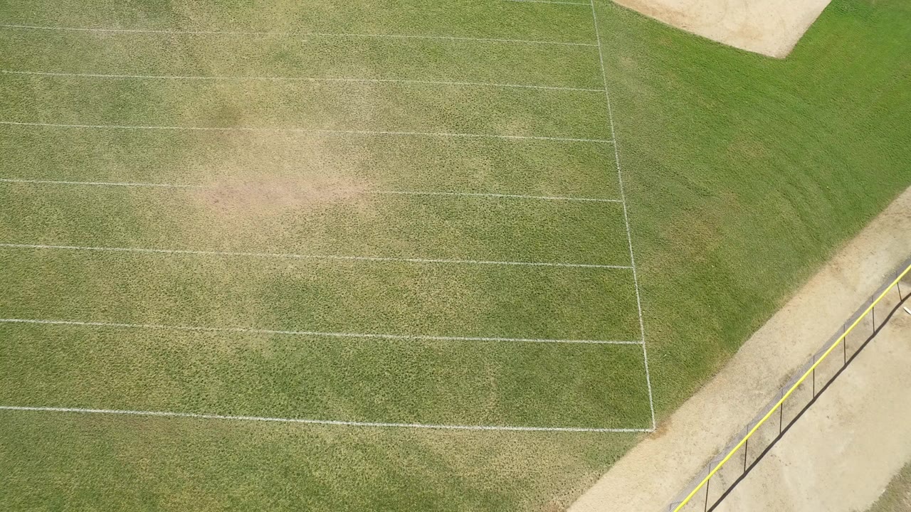 Overhead frame in Hidden Lake, Colorado showing an empty green sports field with white lines, tan worn patches, a paved edge, and no visible people.