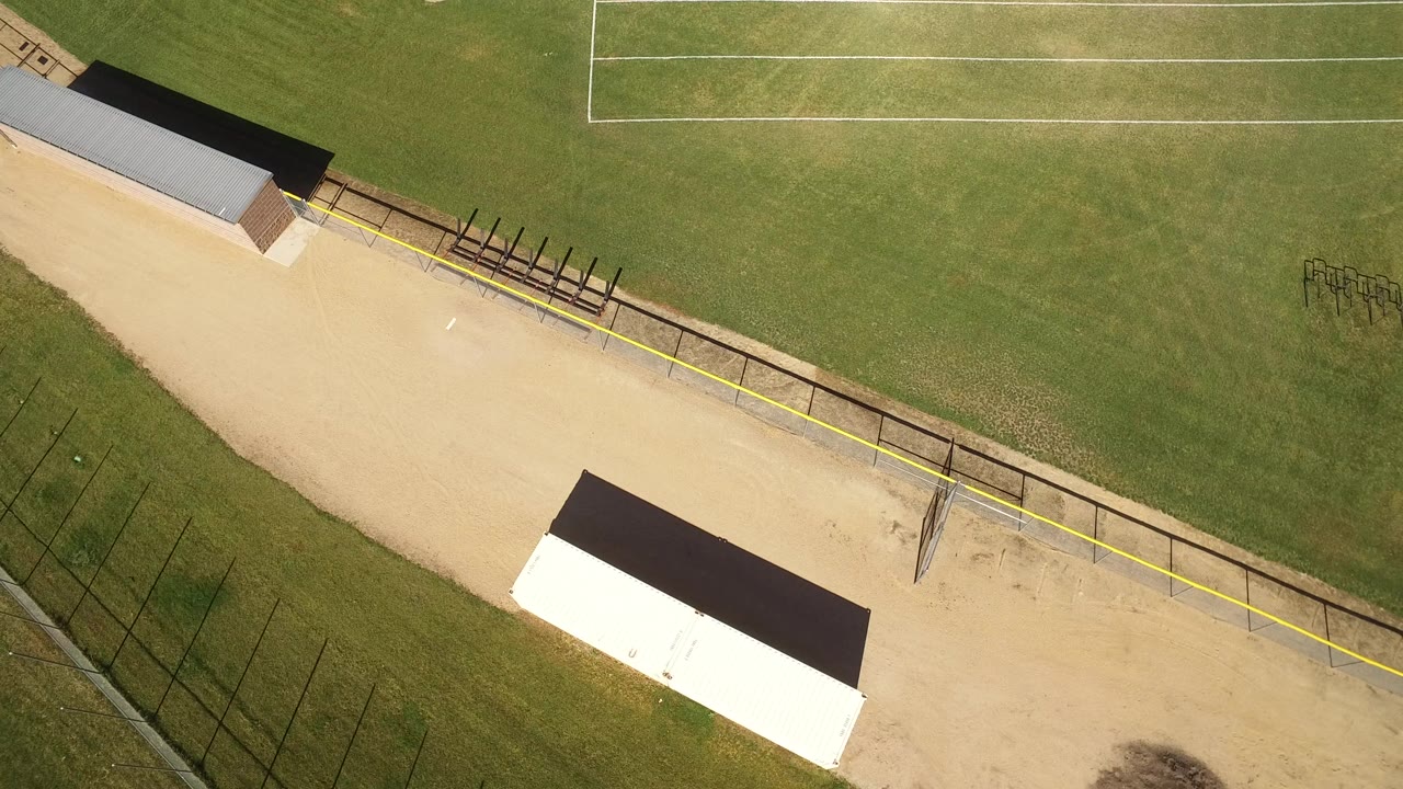 Top-down view of grass, white field lines, dirt path, fencing, and roofed structures.