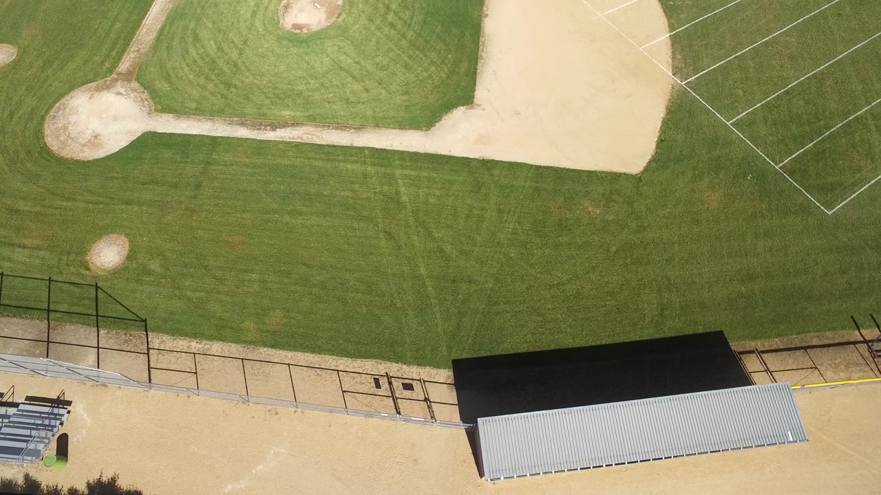 Aerial frame over the Hidden Lake baseball field showing outfield grass, a dirt path to the mound area, fence lines, bleachers, and no visible people.