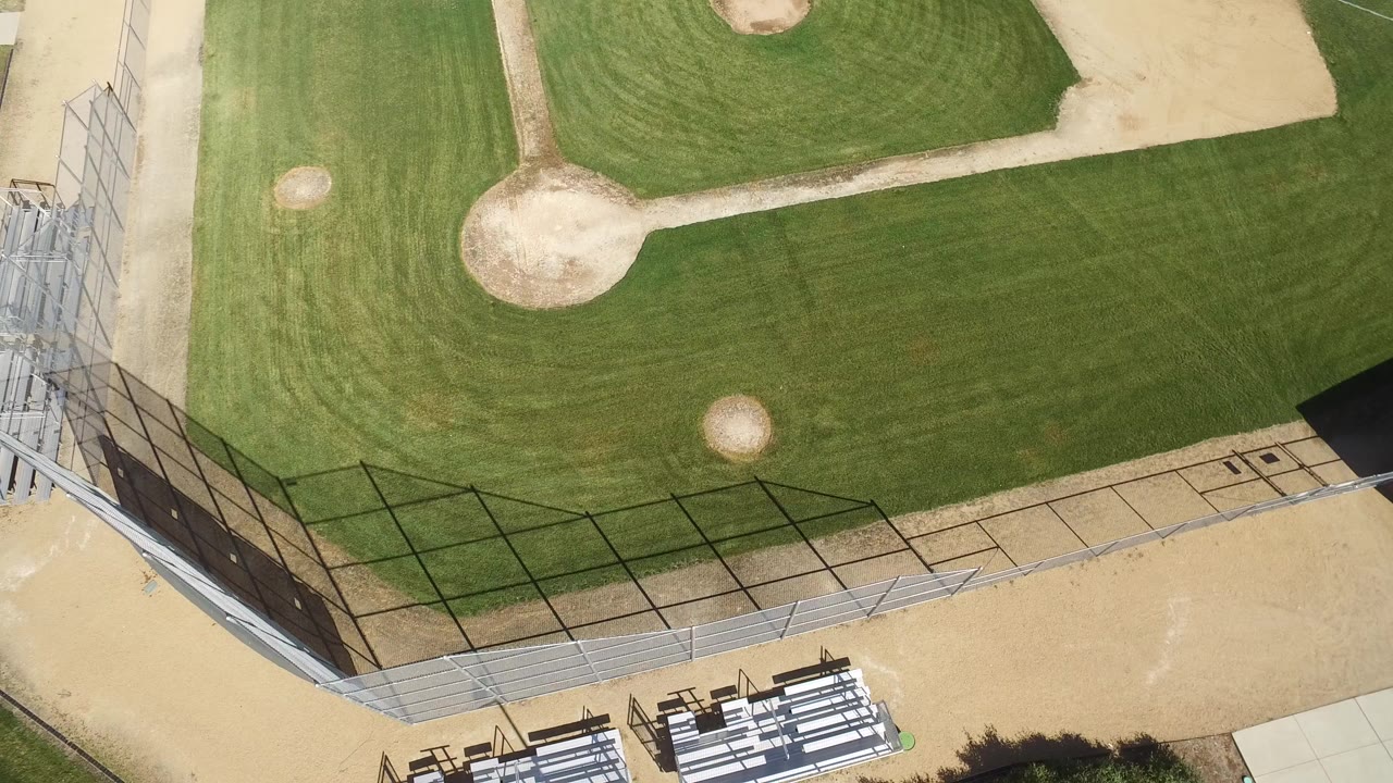 Top-down aerial view of a baseball field with grass, dirt paths, fencing, and bleachers.