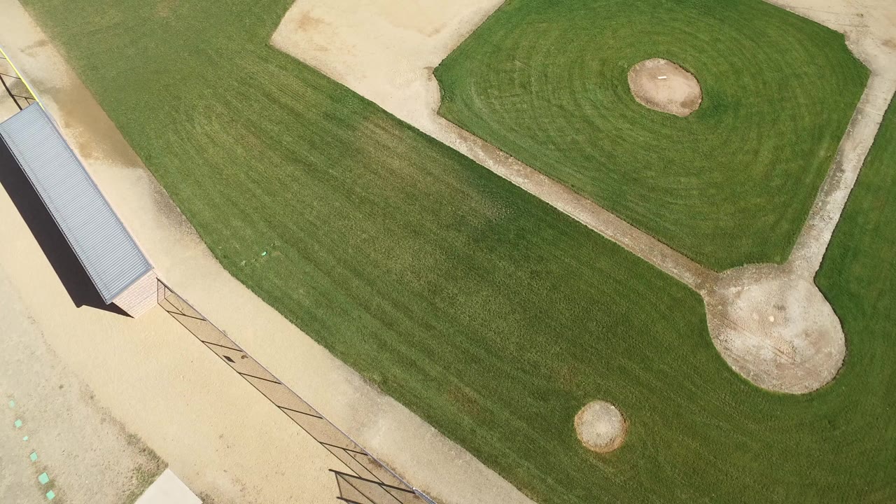 Overhead aerial frame of an empty baseball field in Hidden Lake, Colorado, with the diamond near the top right, a metal-roofed shelter along the left edge, mowed grass, pale dirt, and no visible people.
