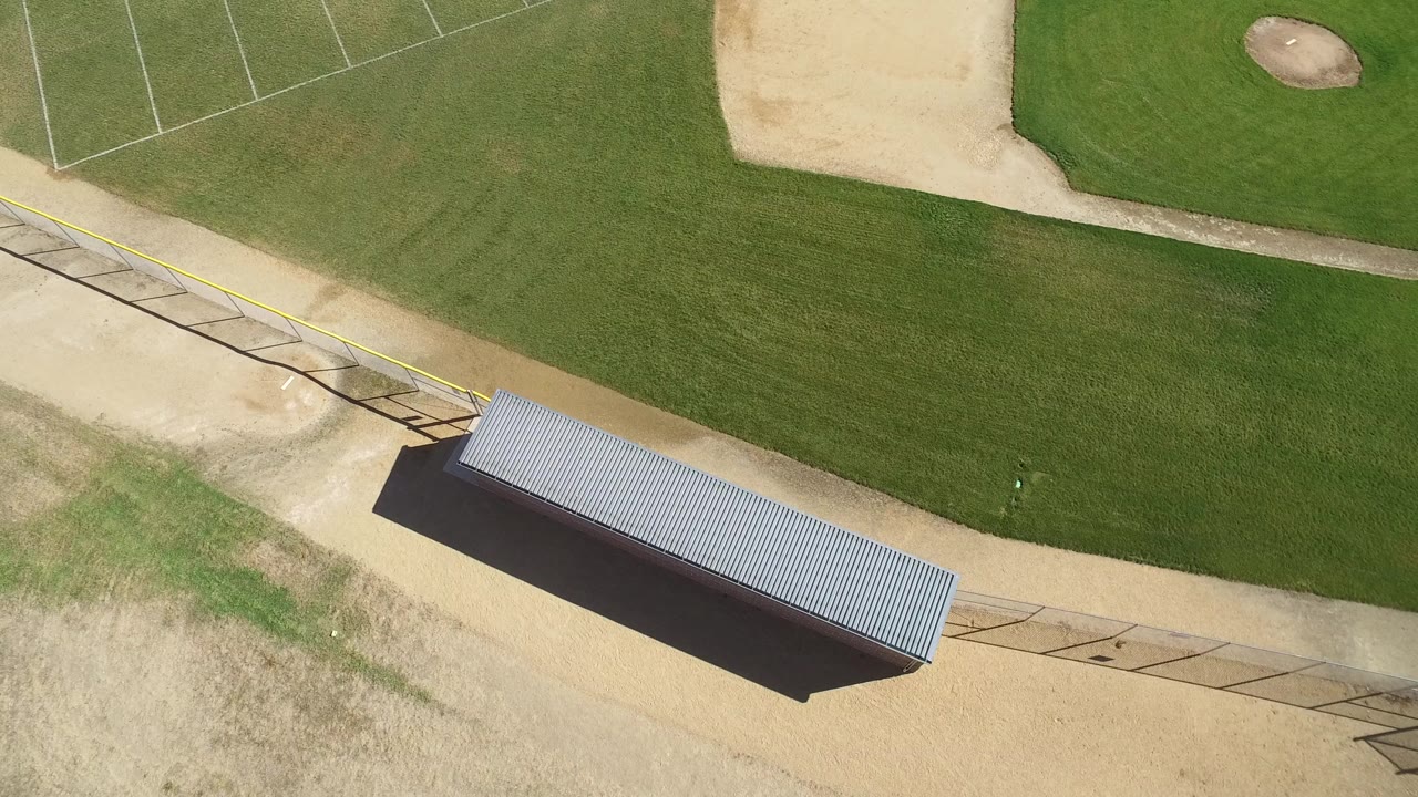 High aerial frame over a Hidden Lake, Colorado baseball field edge, with green grass at top, tan dirt paths, diagonal fencing, a metal-roofed shelter near the bottom, and no visible people.